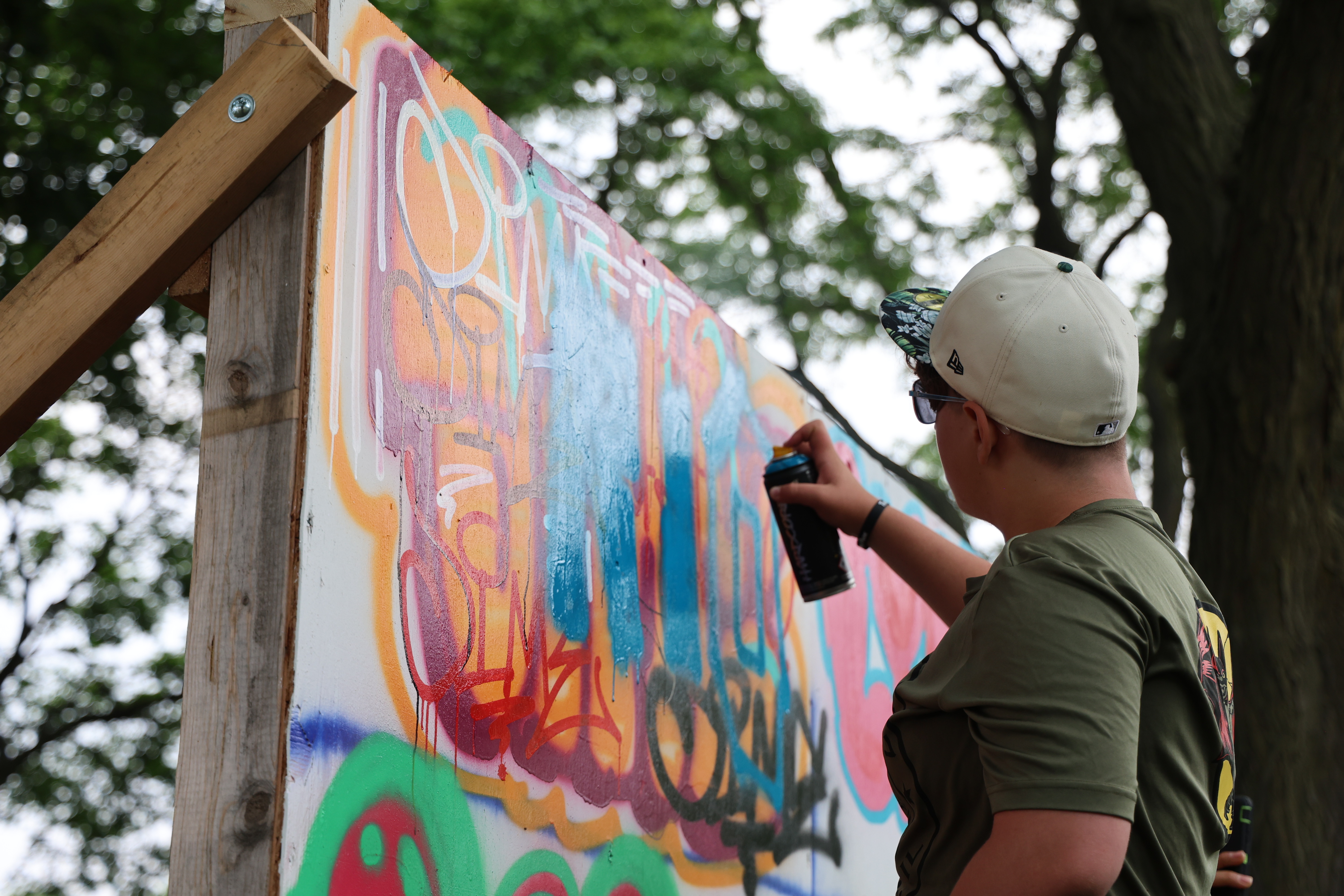 Person adds light blue spray paint to colorful graffiti art on a wooden board.